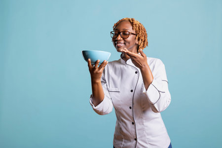Female culinary expert posing against blue background, gently holding bowl of freshly prepared meal. Confident chef with eyes closed and satisfied smile, appreciating aroma of cooked food.の写真素材