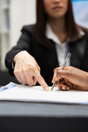 Female receptionist points at clipboard while assisting black patient signing medical insurance forms at hospital front desk. Healthcare staff member provides guidance with registration and paperwork.の写真素材