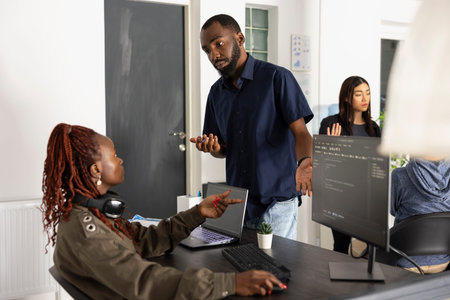 Black male and female developers reviews HTML source code on pc monitor and laptop during meeting. Coworkers communicate ideas clearly while building digital solutions in modern office.の写真素材