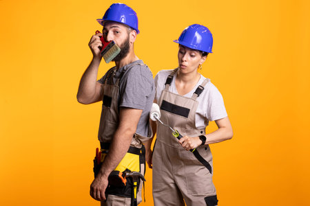 Maintenance crew taking break, woman playfully rolls paint on back of colleague as man holds brush to beard, creating humorous moment at building site. Couple portraying funny moment during diy tasks.の写真素材