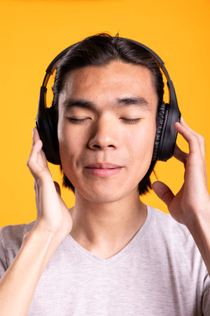 Asian young man listens to peaceful sounds through wireless headset while standing with eyes closed....