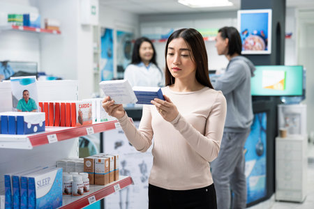 Female customer carefully compares over the counter medicine while standing in drugstore aisle. Adult woman reads product labels and information to ensure she selects correct medication for her needs.の写真素材