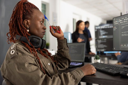 African american woman programmer deep in thought at her workstation, pen at temple, surrounded by multiple screens and colleagues in modern collaborative tech office workspace.の写真素材
