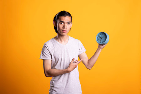 Asian guy gesturing at clock and expressing concern about time. Tensed man looks directly at camera,...