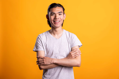 Smiling young Asian man stands confidently with folded arms, showing happiness and fulfillment....