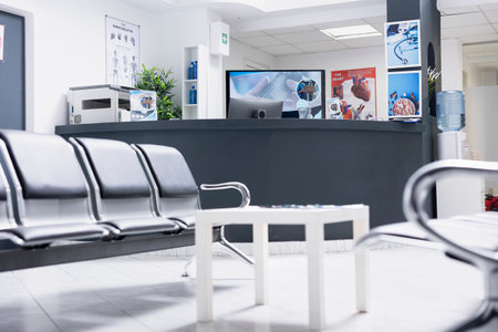Clinical waiting room with vacant chairs, front desk counter, and computer setup ready for hospital...
