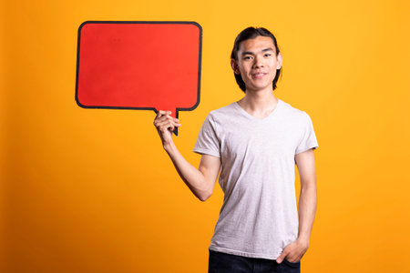 Young asian man stands confidently holding blank placard while posing with one hand in pocket....