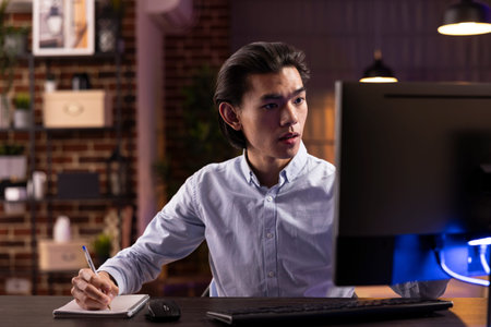 Self employed asian man studying online late at night at home office, using desktop computer for...