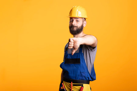Disappointed male contractor signals disapproval with thumbs down. Man wearing protective gear...