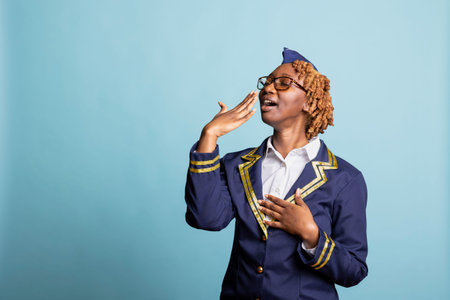 Tired female flight attendant in uniform yawning and covering mouth against blue background. Black...