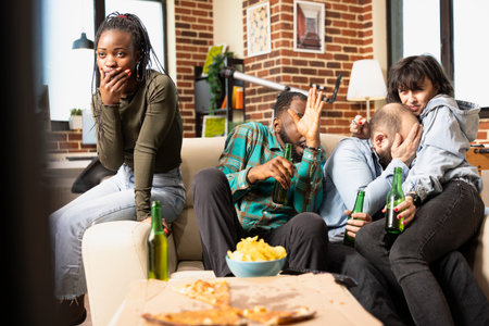 Group of friends seated on couch, holding beer bottles and reacting with terrified expressions....
