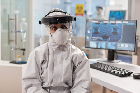 Tired microbiologist in coverall sitting in laboratory looking at camera. Team of scientits...