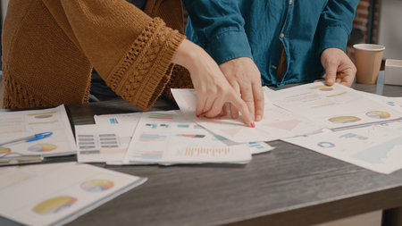 Close up of people checking papers with data rate charts on table. Workmates looking at files to...