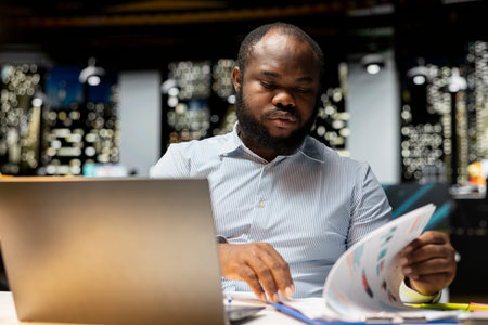 Black advisor researching numbers and finance figures in a dark office at midnight, documenting...