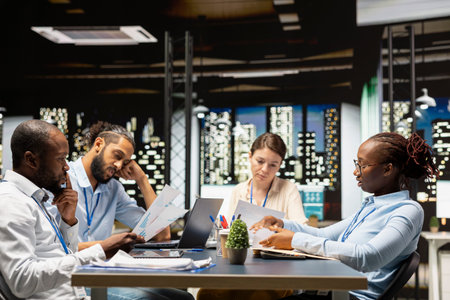 Diverse team of employees in office looking over paperwork documents late at night. Coworkers at...