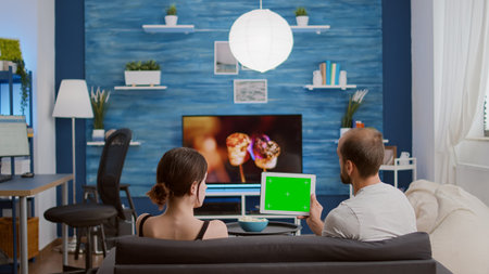 Couple on sofa watching online show using streaming app on digital tablet with green screen in...
