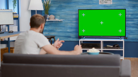 Man sitting on sofa watching sport game on green screen tv mockup encouraging favourite team while...
