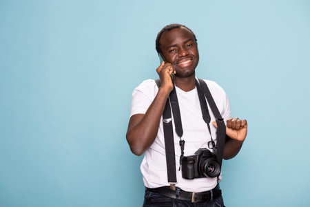 Cheerful black man in suspenders talks on his mobile phone, standing in a blue background studio. He...