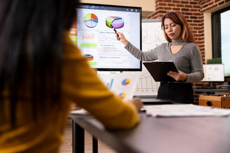 Asian businesswoman reading a document as she points towards the data analytics chart on digital...