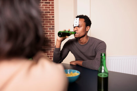 Confident young adult man with plastic card on forehead drinking beer while playing charades with...