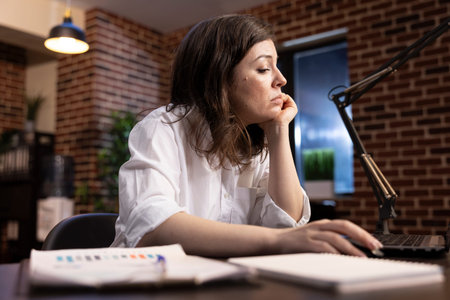 Caucasian female manager working tirelessly at her desk, leaning on one hand while scrolling through...