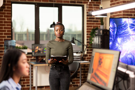 Portrait of african american woman standing in brick wall office and holding a clipboard. Confident...