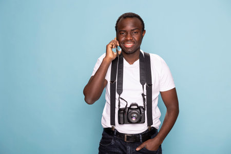 Smiling black man looking at the camera with one hand in his pocket while answering a call on his...