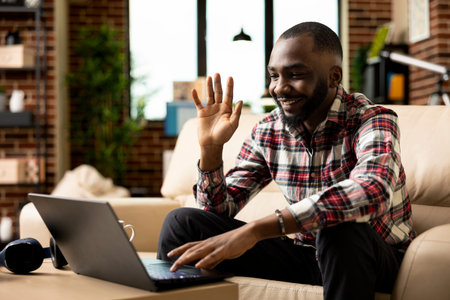 Happy self employed man seated on sofa, waving at device screen, attending online business course....