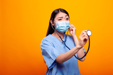 Young female medic wearing face mask and holding a stethoscope, standing against a yellow background. Asian woman in a nurse uniform grasping a medical equipment in front of an isolated backdrop.の写真素材