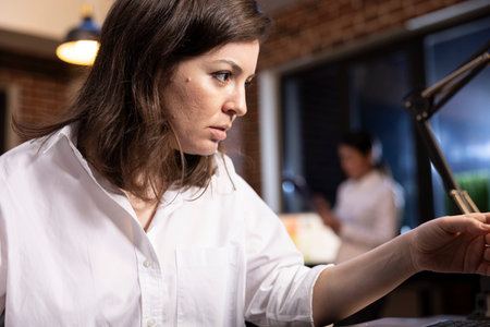A determined woman in a white shirt works late at her office desk, deeply focused on her task. A...