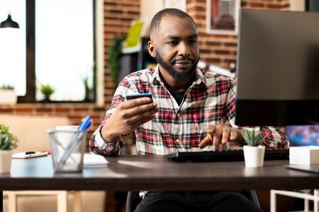 Bearded male freelancer seated at computer, purchasing item online with credit card in hand, making...