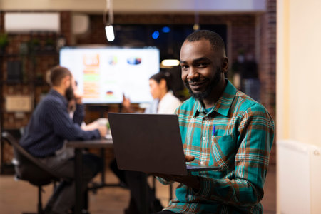 Professional man seated with laptop in modern workspace, looks at camera while working late. Male...