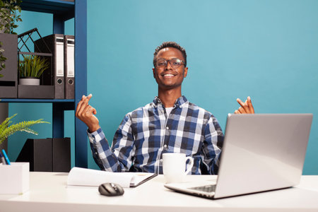 Smiling young man gesturing for cash by rubbing his fingers in a playful payday moment on a blue...
