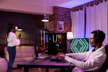 Gamer immersed in gaming session at computer desk while girlfriend in background uses virtual...