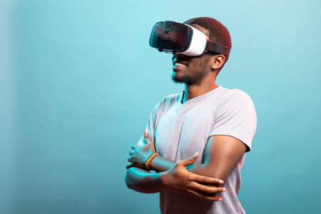 Young male individual enjoying an immersive VR experience, interacting with a 3D simulation. Portrait of african american tech enthusiast wearing virtual reality goggles, stands with crossed arms.の写真素材