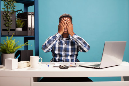 Frustrated male consultant sits at office desk with a laptop and coffee, feeling overwhelmed. Tired...