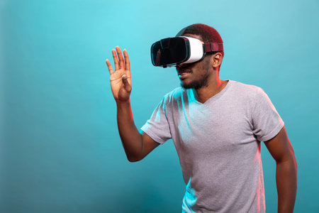 Young male adult using VR goggles, exploring a 3D digital world and engaging with interactive technology. Black tech enthusiast immersed in futuristic simulation, trying out a virtual reality device.の写真素材