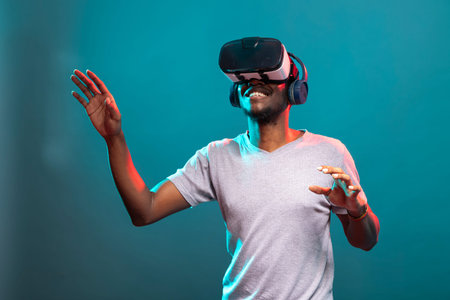 Cheerful young man standing over blue background and experiencing an interactive 3D virtual...