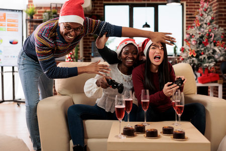 Cheerful employees in santa hats laughing and playing console game on christmas in decorated office....