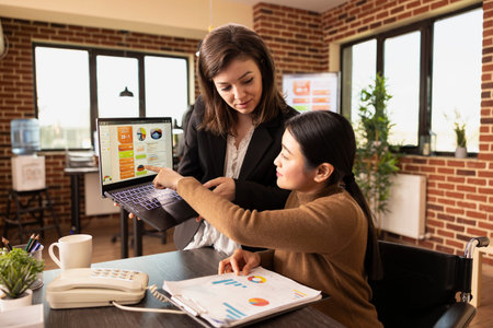 Caucasian employee stands holding laptop, receiving guidance from her asian manager in brick wall...