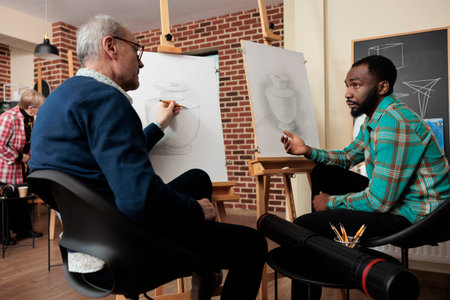 Two diverse men art school students talking during lesson, sitting at easels discussing artwork...