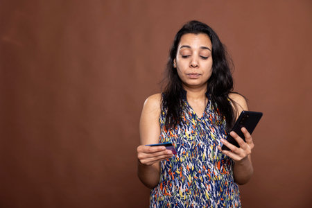 Confident female individual using mobile device to complete online transaction in brown background studio. Indian woman holding smartphone and debit card, completing secure digital payment.の写真素材