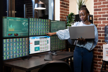 African American stockbroker standing by desk, comparing forex market data on laptop and several...
