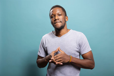 Portrait of african american person in casual t shirt, standing with fingers clasped against blue...