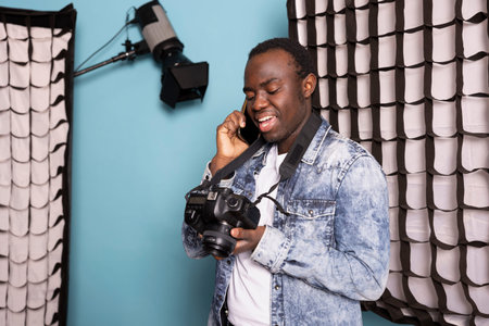 Black photographer on phone call, holding his DSLR and standing near his studio equipment. African...