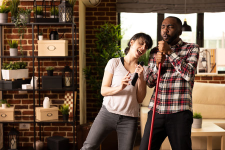 Playful husband and wife cleaning together in living room, with white woman using tv remote as...