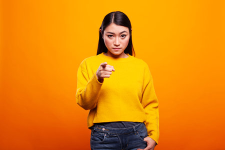Serious woman in a yellow sweater pointing directly at the camera on an orange background. Young asian lady standing with a stern look and gesturing her index finger towards the viewer.の写真素材