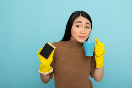 Pretty female cleaner wearing yellow rubber gloves holds coffee mug to mouth, taking break from...