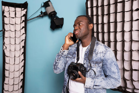 Professional male photographer wearing denim jacket holding his camera in one hand and talking on...