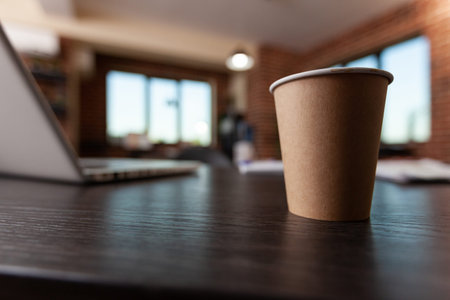 Close up of plastic coffee cup on wooden desk with laptop used for business work in empty startup...